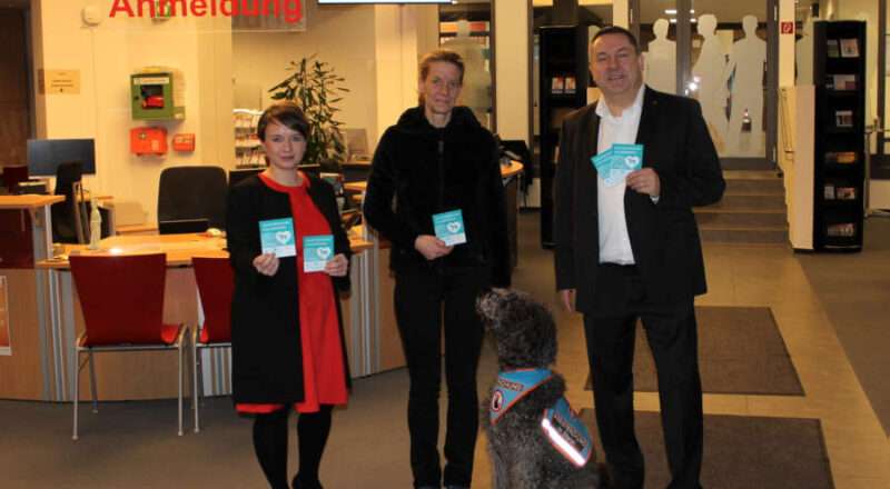 Gelebte Inklusion: Kreismitarbeiterin Diana Helms (Mitte) freut sich, dass ihr Assistenzhund „Fix“ im Kreishaus willkommen ist. Gemeinsam mit Landrat Dr. Axel Lehmann und Inklusionsbeauftragter Monika Heel bringt sie die ersten Aufkleber der Kampagne im Bürgerservice an. (Foto: Kreis Lippe)