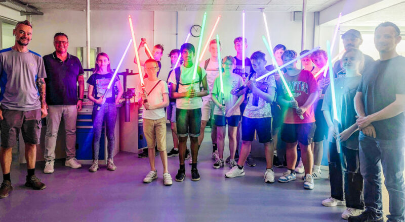 Große Lichtschwertshow in der abgedunkelten Metallwerkstatt: Von links: Dozent Kai Lenski (Argentumsabers), Thomas Mahlmann (zdi-Zentrum Lippe.MINT), links: Dozent Erwin Schadt (Metallwerk OWL/Lippe Bildung eG), dritter von links: Dozent Lukas Dusche (Institut für die Kunststoffwirtschaft/Lippe Bildung eG) mit den stolzen Schülerinnen und Schülern des Lichtschwertkurses. Foto: Lippe Bildung eG