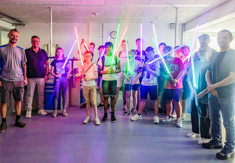 Große Lichtschwertshow in der abgedunkelten Metallwerkstatt: Von links: Dozent Kai Lenski (Argentumsabers), Thomas Mahlmann (zdi-Zentrum Lippe.MINT), links: Dozent Erwin Schadt (Metallwerk OWL/Lippe Bildung eG), dritter von links: Dozent Lukas Dusche (Institut für die Kunststoffwirtschaft/Lippe Bildung eG) mit den stolzen Schülerinnen und Schülern des Lichtschwertkurses. Foto: Lippe Bildung eG