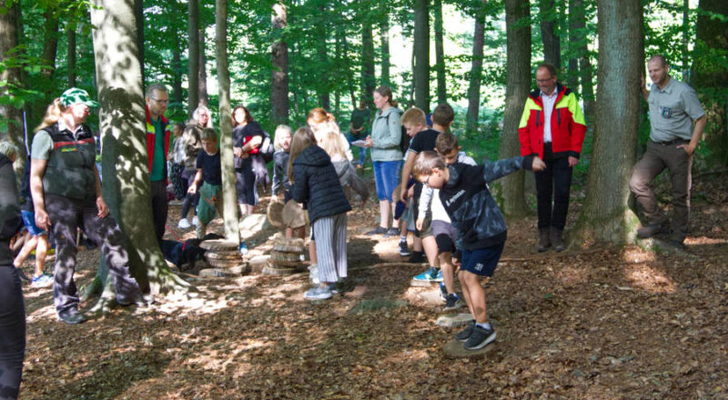 Kinder der vierten Klasse probieren in Teams, so schnell wie möglich mit Holzscheiben über das „Moor“ zu kommen. Johanna Ahlmeier (Regionalforstamt Ostwestfalen-Lippe, l.), Jan-Otto Hake (Leiter Forstabteilung Landesverband Lippe, 2.v.l.), Jörg Düning-Gast (Verbandsvorsteher Landesverband Lippe, 2.v.r.) und Thomas Schulte (Leiter Forstbetriebsbezirk Detmold und Organisator der Waldjugendspiele, r.) schauen den Kindern zu. Foto: Landesverband Lippe