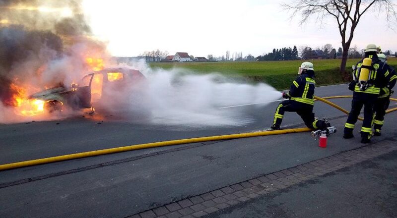 Ein Feuerwehrmann aus sicherem Abstand Wasser auf das brennende Auto, während zwei Atemschutzgeräteträger sich auf ihren Einsatz vorbereiten. Foto: Freiwillige Feuerwehr Leopoldshöhe