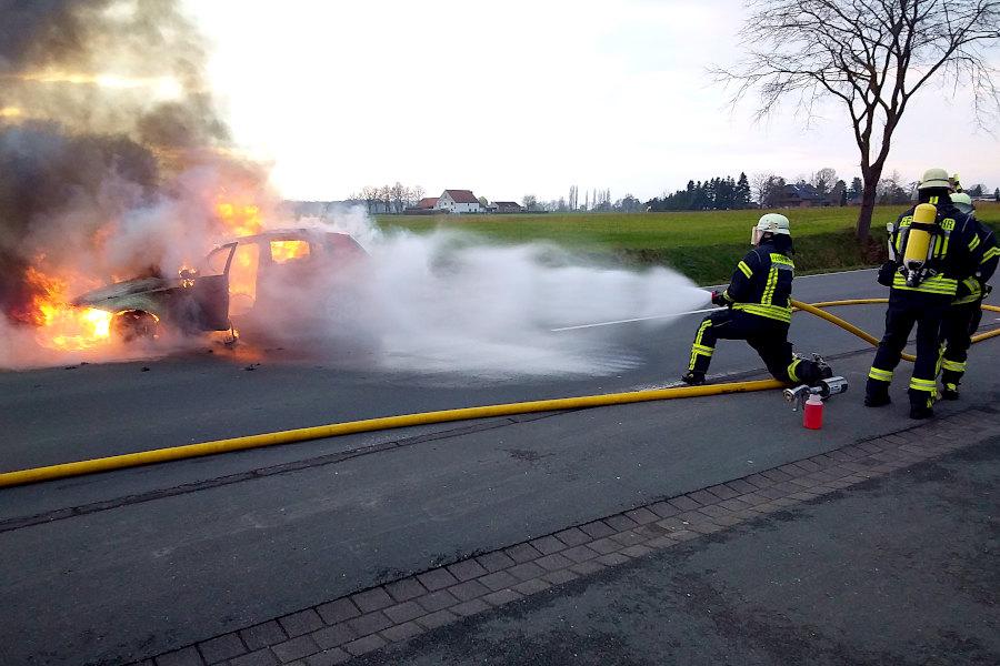 Ein Feuerwehrmann spritzt aus sicherem Abstand Wasser auf das brennende Auto, während zwei Atemschutzgeräteträger sich auf ihren Einsatz vorbereiten. Foto: Freiwillige Feuerwehr Leopoldshöhe