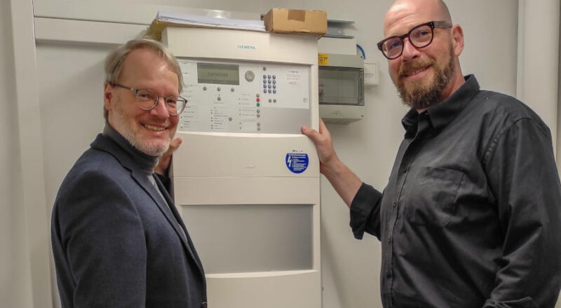 Dr. Joachim Eberhardt (Direktor der Lippischen Landesbibliothek / Theologischen Bibliothek und Mediothek Detmold, l.) und Andreas Prange (Architekt, Infrastrukturabteilung Landesverband Lippe) vor der Brandmeldezentrale – das Herzstück der Brandmeldeanlage. Foto: LLB