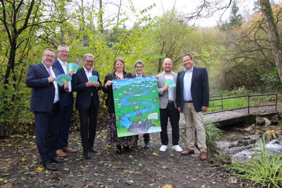 Freuen sich vor einer Wasser-im-Fluss-Maßnahme an der Berlebecke über den Geburtstag des Projektes (von links): Landrat Dr. Axel Lehmann; Dr. Nicolas Rinösl, Abteilungsleiter für Umwelt und Arbeitsschutz bei der Bezirksregierung Detmold; Dr. Lucas Heumann, Vorstandsvorsitzender der AGA gGmbH; Jennifer Strunk, Projektplanerin bei der AGA gGmbH; Jürgen Benning, Projektverantwortlicher beim Kreis Lippe; Robin Debuisson, Projektleiter bei der AGA gGmbH und Jens Fillies, Geschäftsführer der AGA gGmbH. Eine eigens für das Jubiläum gestaltete Illustration zeigt die Entwicklung des Projektes. Foto: Kreis Lippe