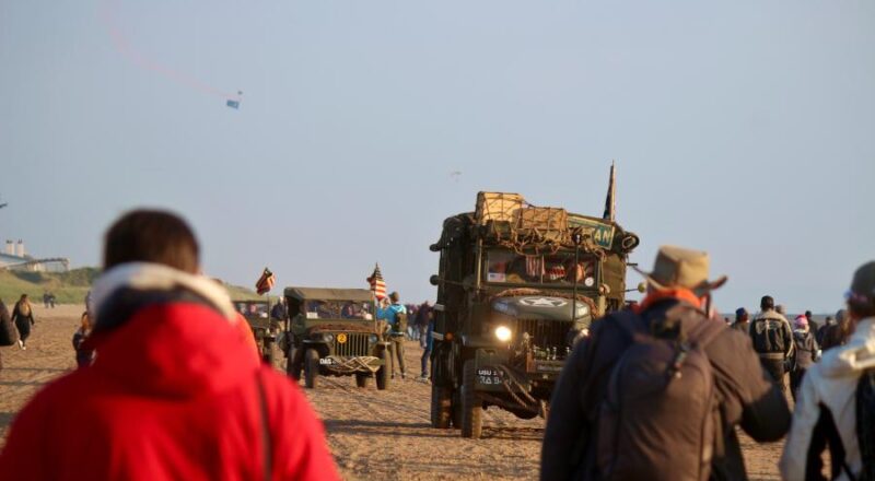 Historische Militärfahrzeuge fahren am Strand entlang, während im Hintergrund am Himmel US-Navy Seals zu Boden segeln. Foto: Thomas Dohna