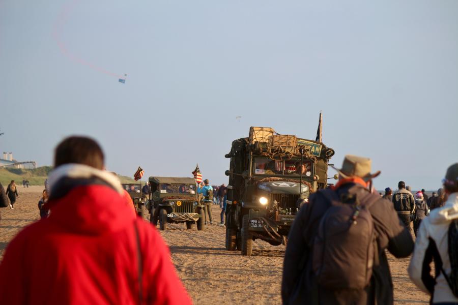 Historische Militärfahrzeuge fahren am Strand entlang, während im Hintergrund am Himmel US-Navy Seals zu Boden segeln. Foto: Thomas Dohna