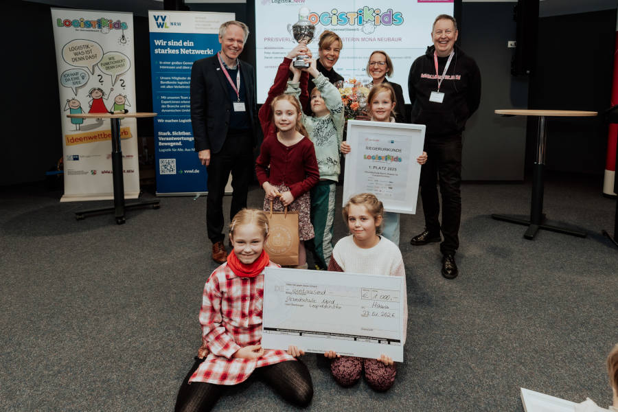 Sind stolz auf ihre Urkunde und den Scheck – vorne sind die Kinder der Grundschule Nord aus Leopoldshöhe. Dahinter: Ocke Hamann, Mona Neubaur, Madlen Klarhorst, Moderator Peter Abelmann (von links). Foto: Nico Schmitz/Kompetenznetz Logistik.NRW