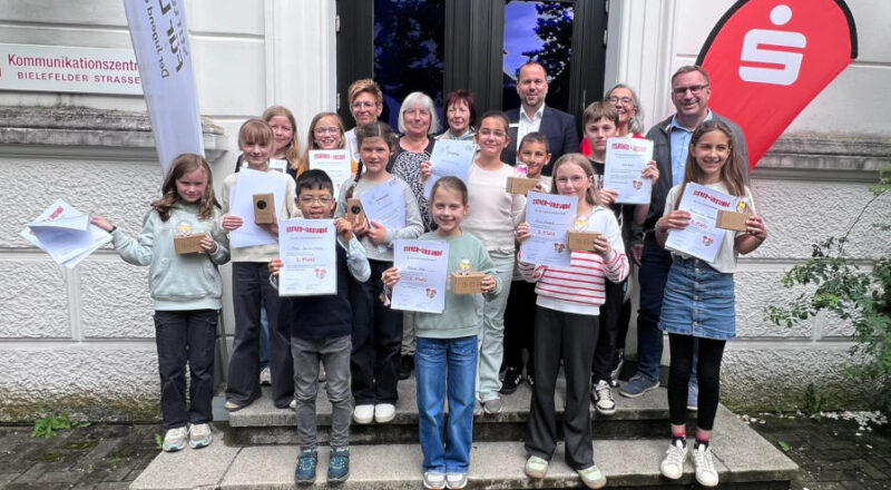 Oscar Squiccimarro (vorne, von links), Marie Eke, Elin Schwarz und Minou Lea Tawalalli belegten beim Vorlesewettbewerb der lippischen Grundschulen die ersten Plätze. Mit ihnen freuten sich die weiteren Finalistinnen und Finalisten sowie die Jury vor dem Kommunikationszentrum der Sparkasse Paderborn-Detmold-Höxter. Foto: Dr. A. Heinrike Heil/ Stiftung Standortsicherung