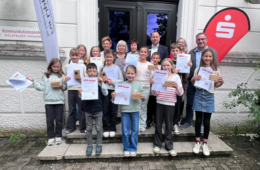 Oscar Squiccimarro (vorne, von links), Marie Eke, Elin Schwarz und Minou Lea Tawalalli belegten beim Vorlesewettbewerb der lippischen Grundschulen die ersten Plätze. Mit ihnen freuten sich die weiteren Finalistinnen und Finalisten sowie die Jury vor dem Kommunikationszentrum der Sparkasse Paderborn-Detmold-Höxter. Foto: Dr. A. Heinrike Heil/ Stiftung Standortsicherung